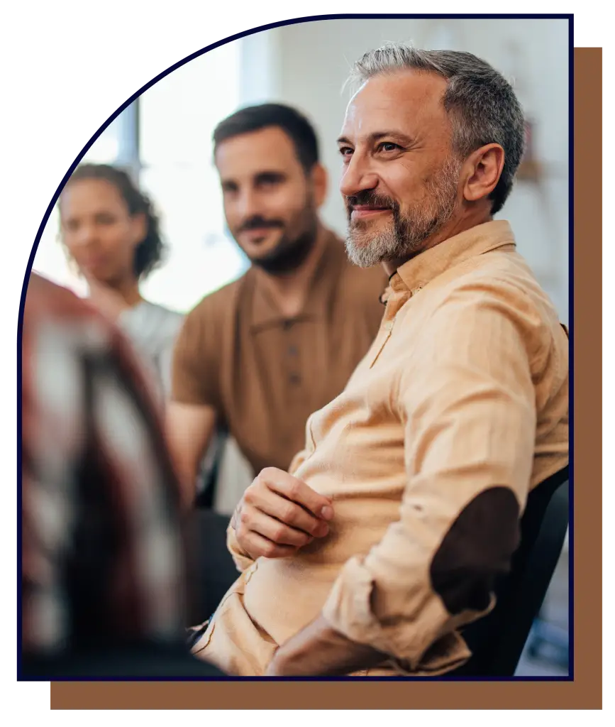 A man with grey hair and a beard sitting in a group setting listening to someone talk