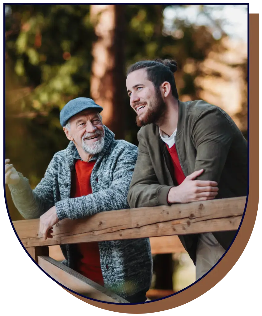An older man with a beard and a hat, having a conversation with a younger man with a beard, while both are leaning on a wooden railing, they are both smiling or laughing