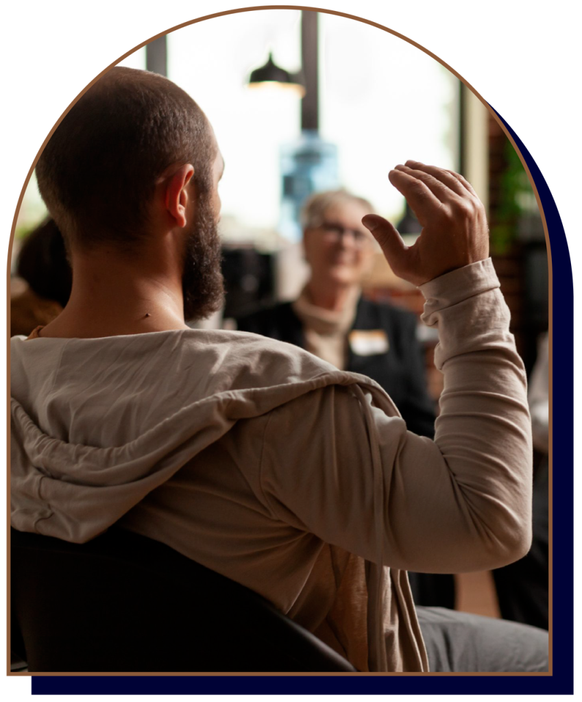 Man with beard gesturing while speaking to others indoors.